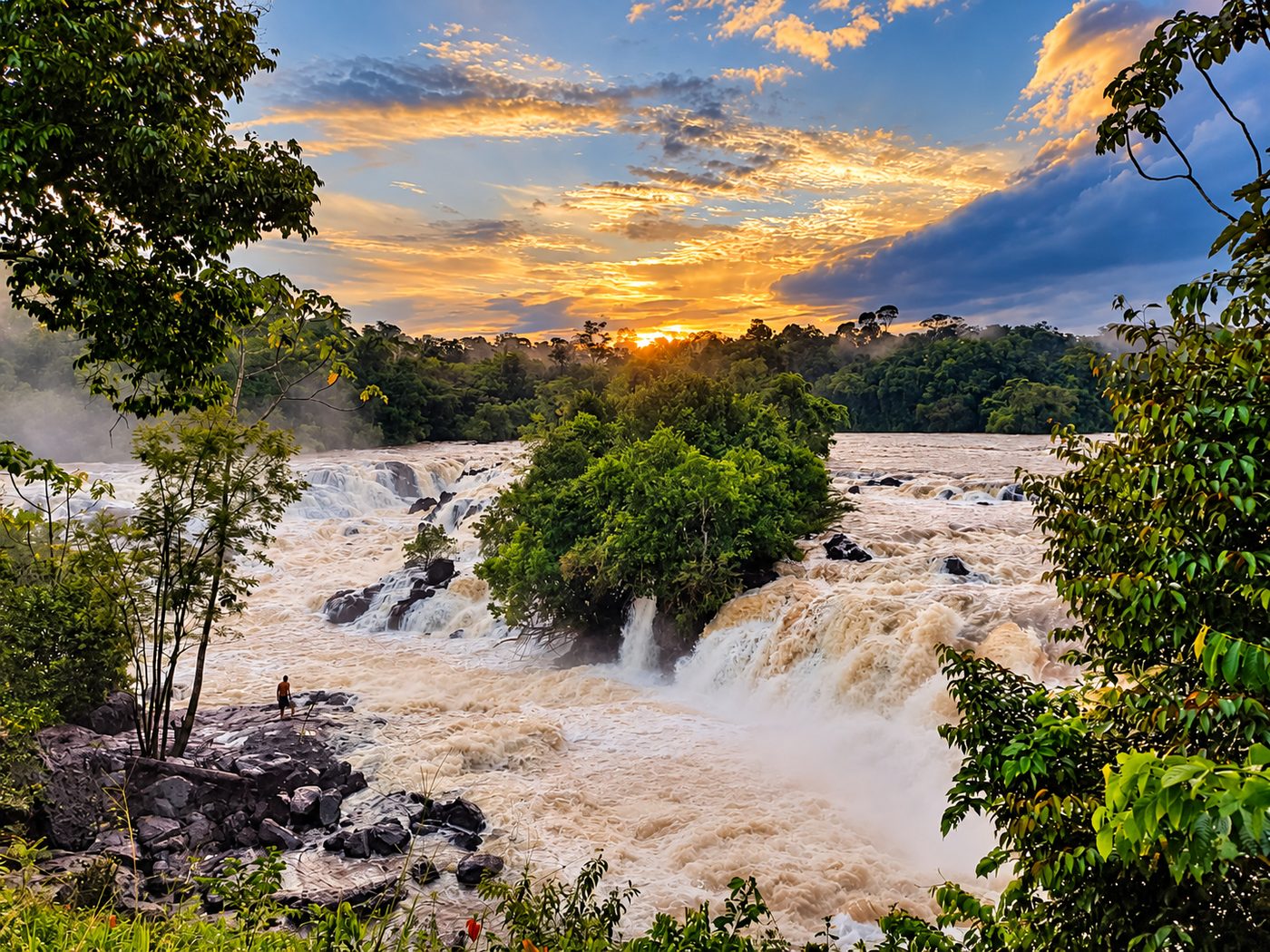 The Blanche-Marie falls at sunset: wide rapids pour around a forested island, with a lone visitor standing on the rocks in the foreground.